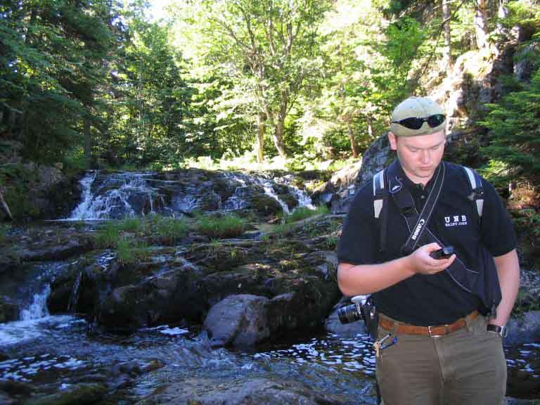 Photographie couleur d’un ruisseau, d’arbres et de roches et d’un homme avec GPS