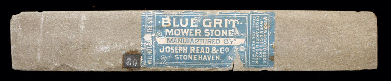 Photographie couleur d’une pierre grise allongée avec une étiquette « Blue Grit Mower Stone » (pierre à affûter en grès grossier bleu)
