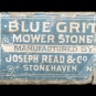 Photographie couleur d’une pierre grise allongée avec une étiquette « Blue Grit Mower Stone » (pierre à affûter en grès grossier bleu)
