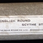 Photographie couleur d’une pierre grise allongée avec une étiquette « English Round Scythe Stone » (pierre ronde anglaise à faux)