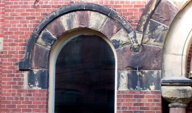 Photographie couleur d’une porte voûtée entourée de brique rouge et surmontée de grès rouge et brun