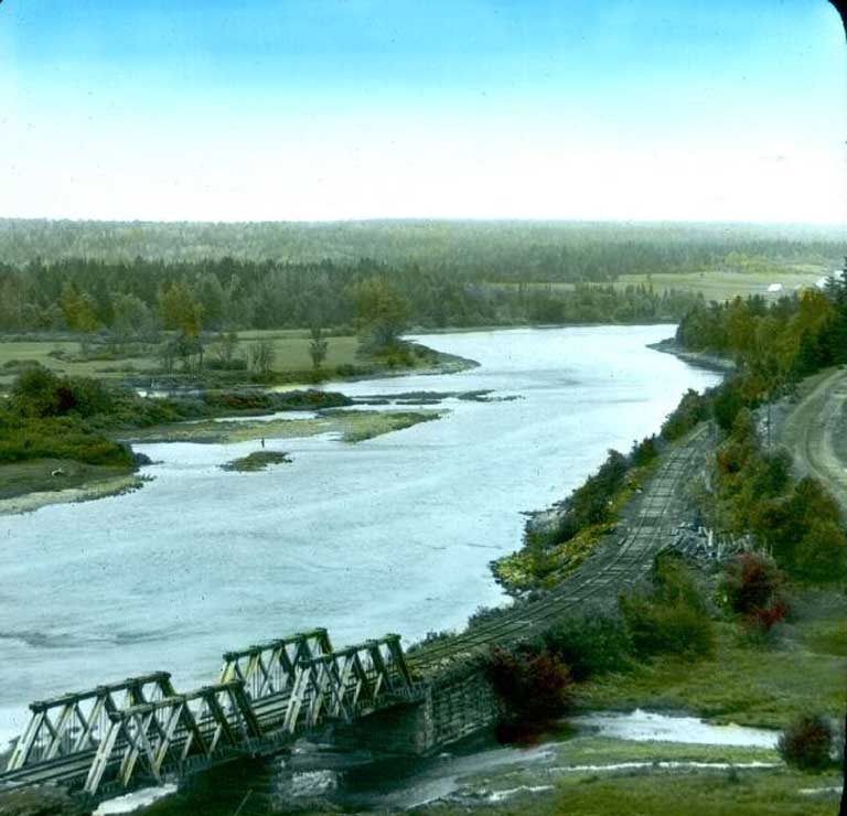 Photographie couleur d’un cours d’eau entouré d’arbres et de champs verdoyants, qui longe un pont ferroviaire