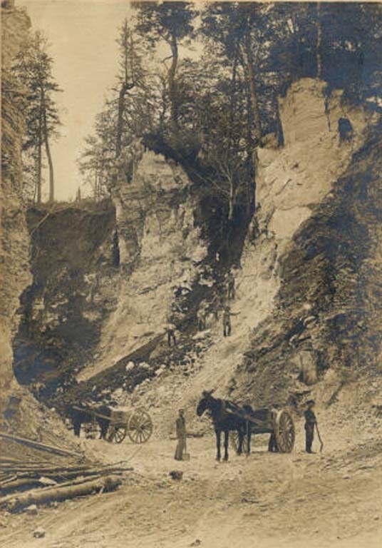 Black and white photograph of steep cliffs, two horses with wagons and several men with shovels
