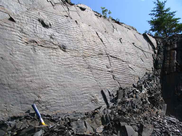 Colour photograph of gray rock face with horizontal striping and hammer for scale