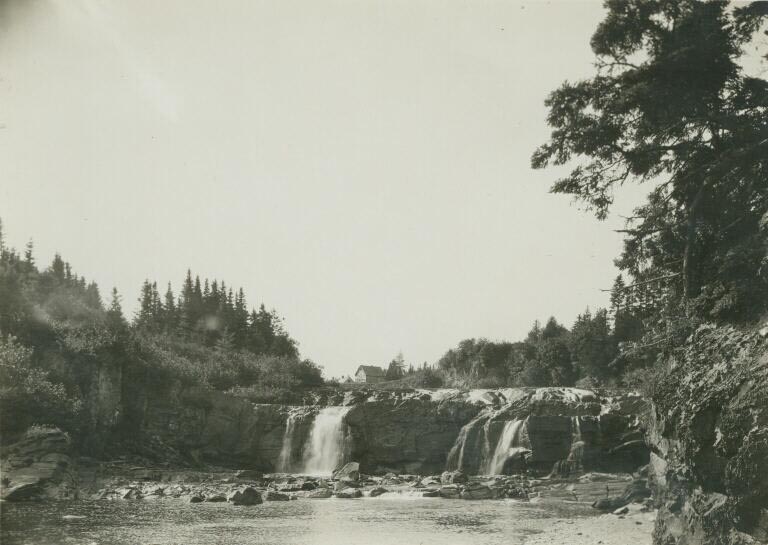Black and white photograph of waterfall and rocky ledges and shoreline in distance