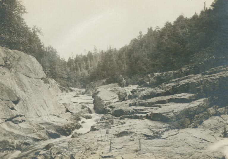 Black and white photograph of rocks along a river with trees along the shore