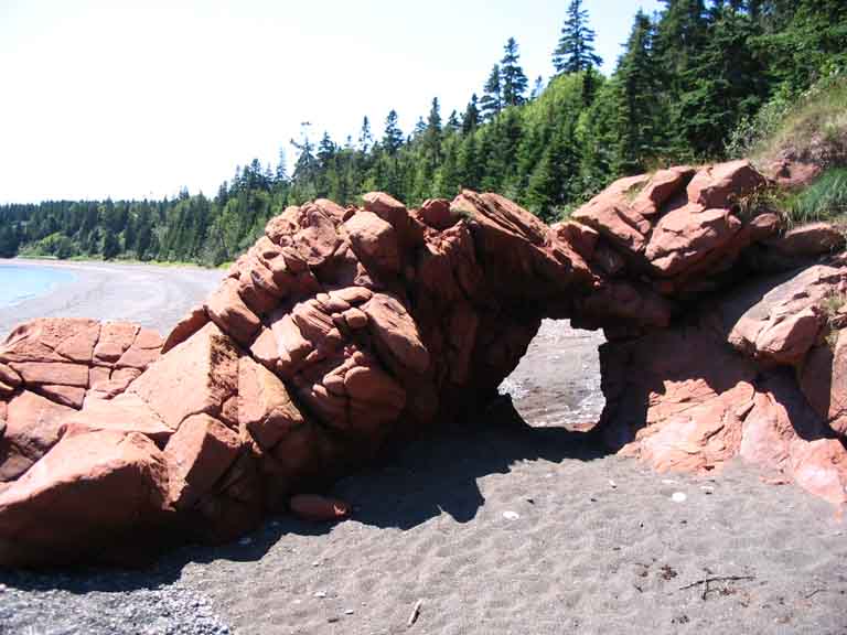 Colour photograph of a red sandstone rock on the ocean with a beach and trees on top of the cliff