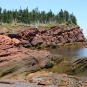 Colour photograph of a red sandstone rock on the ocean with a beach and trees on top of the cliff