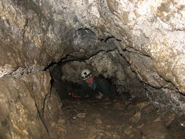 Colour photograph of man in white hard hat sliding through cave