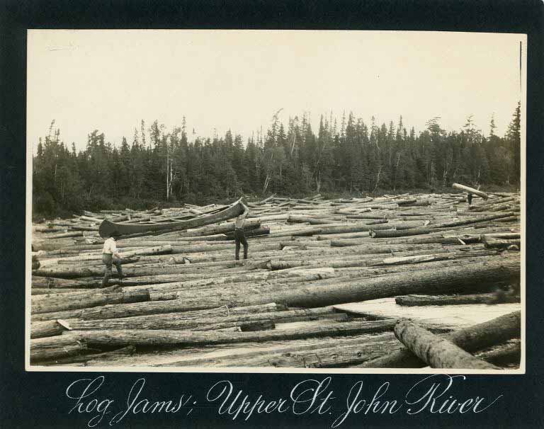 Black and white photograph of three men carrying canoes across logs on a river