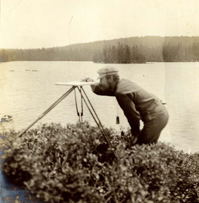Black and white photograph of man standing in front of a lake with surveying equipment
