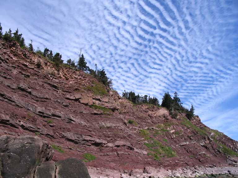 Colour photograph of a cliff of red rocks and a blue sky with clouds