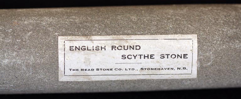 Colour photograph of a long gray stone with a label ‘English Round Scythe Stone’