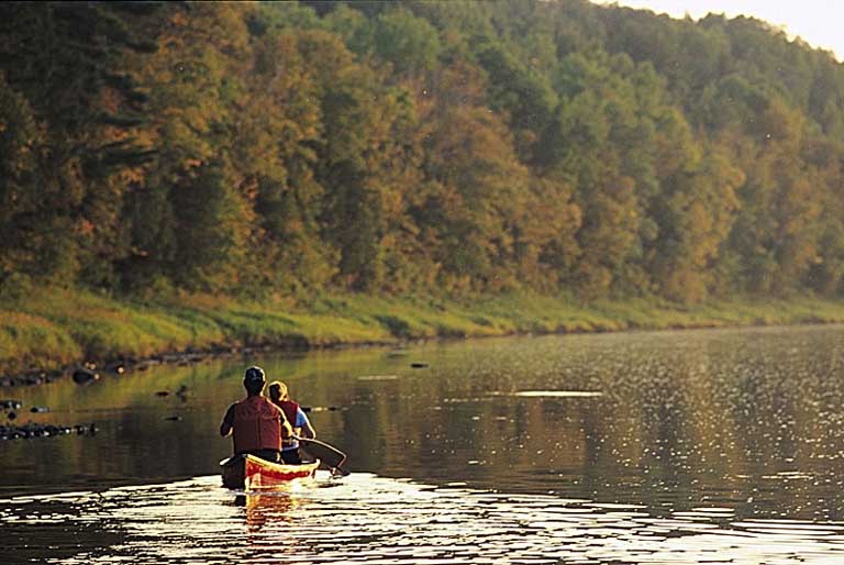 Colour photograph of a river with trees along the shore and two people in a canoe