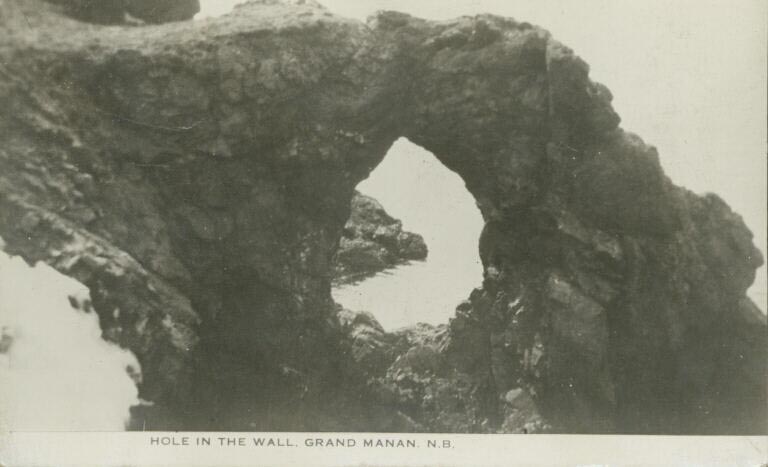 Black and white postcard of curved rock formation with hole in the middle on shoreline