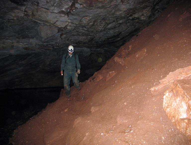 Photographie couleur d’une personne portant combinaison de travail verte et casque de protection blanc debout dans une caverne