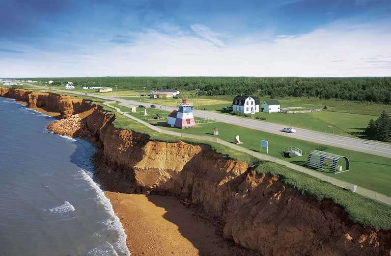 Photographie couleur d’un rivage de falaises brunes avec, au sommet, des champs verdoyants qui entourent le bâtiment d’un phare