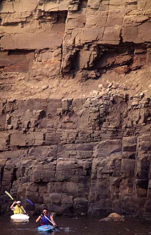Photographie couleur d’une falaise de roche brune au bord de l’océan avec deux personnes dans des kayaks