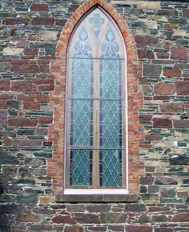 Colour photograph of gray, red and black stone wall and arched, multi-paned church window