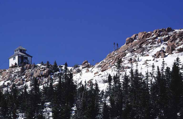 Colour photograph of rocky mountain peak with trees, snow and building