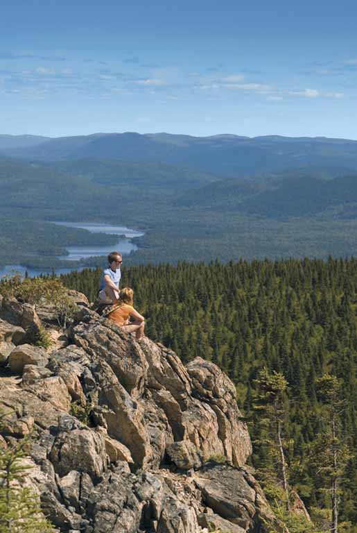 Colour photograph of rocky summit, man, woman overlooking lake, trees, valley and other mountains