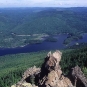 Colour photograph of rocky mountain point overlooking lake, trees, valley and other mountains