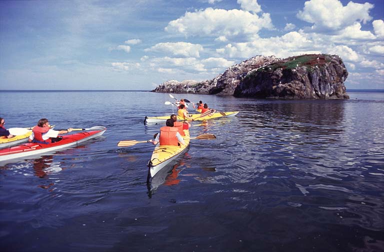 Colour photograph of several kayaks and people approaching a large rock formation