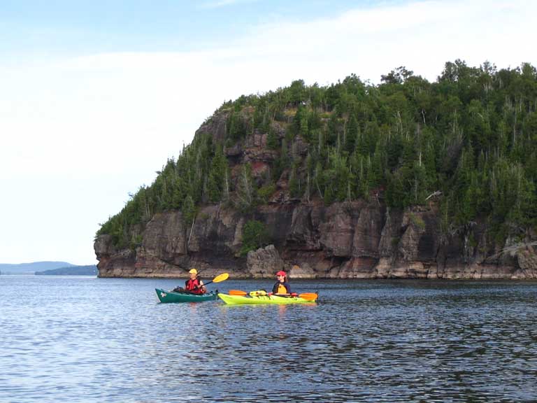 Colour photograph of two kayaks on calm water near rocky peninsula with trees