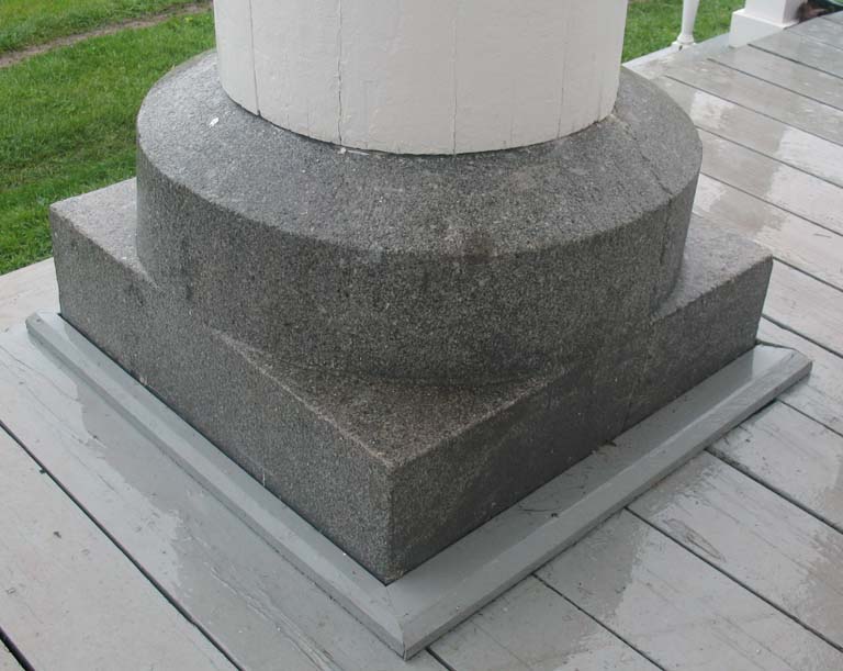 Colour photograph of gray speckled rock carved square at bottom and circular at the top, supporting wooden white column