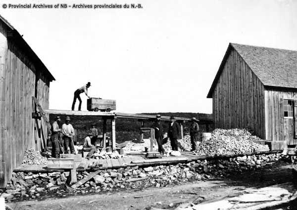 Photographie noir et blanc montrant une mine de roches, des bâtiments, des hangars, des roches et un homme sur le toit d'un hangar