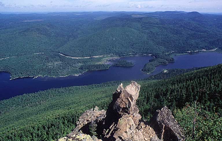 Photographie couleur d’un piton rocheux dominant un lac, des arbres, la vallée et d’autres montagnes