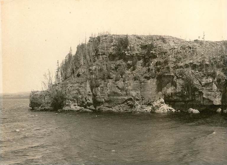Photographie noir et blanc montrant de l’eau et une presqu'île rocheuse couverte d’arbres