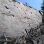 Colour photograph of gray rock face with horizontal striping and hammer for scale