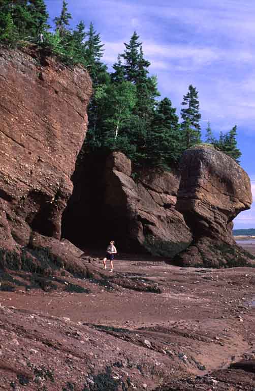 Colour photograph of red rock cliffs and tall formations with person standing on beach