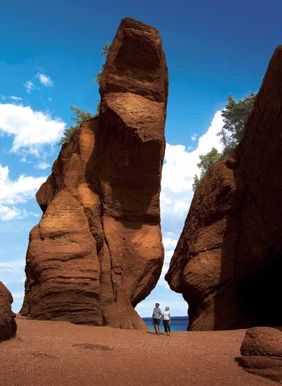 Colour photograph of tall, red rock formation with two people on beach