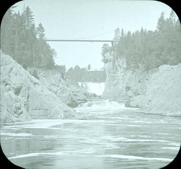 Black and white image of river, rocky shoreline and suspension bridge in the background