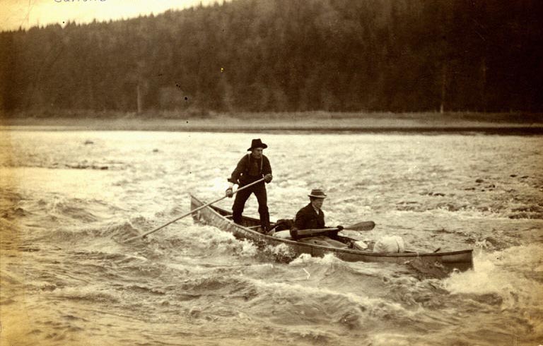 Photographie noir et blanc de deux hommes, dont un debout, en train de descendre des rapides