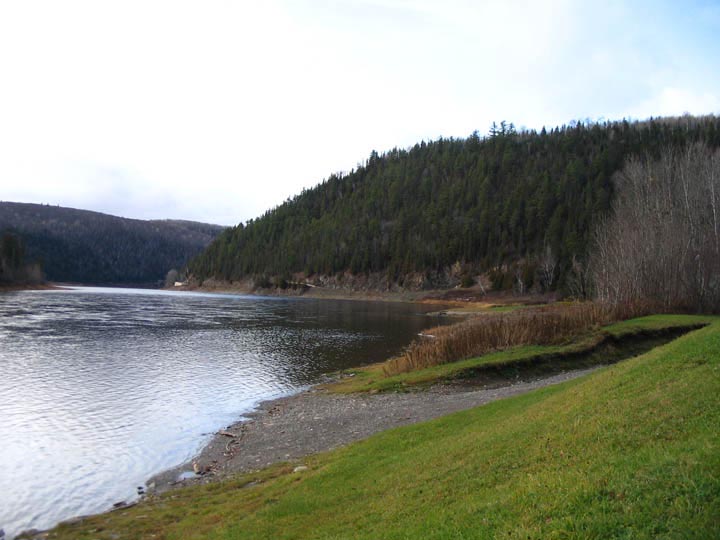 Photographie couleur de la rivière, d’une plage de galets et de collines couvertes d’arbres