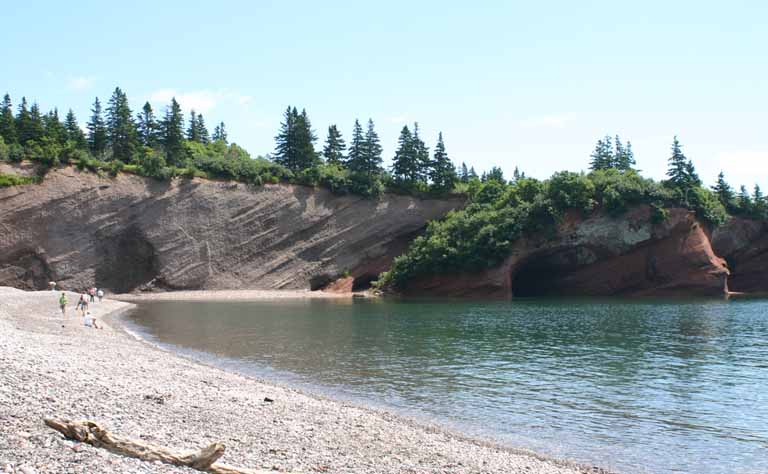 Colour photograph of a red rock cliff on the ocean with a beach and trees on top of the cliff