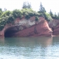 Colour photograph of a red rock cliff on the ocean with trees on top of the cliff