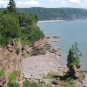 Colour photograph of a red rock cliff on the ocean with a beach and trees on top of the cliff