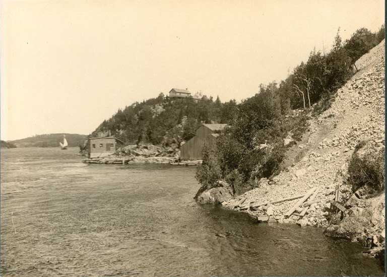 Photographie noir et blanc de cours d’eau, rive rocailleuse et de maisons en arrière-plan