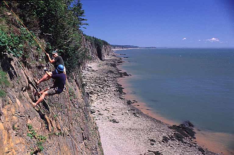 Colour photograph rock cliff along a shoreline and two people rock climbing